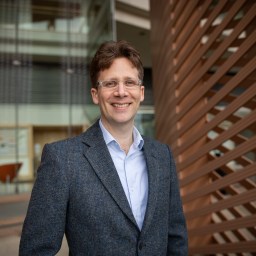 A man wearing glasses, a blue shirt, and a grey blazer stands outdoors, smiling, with a modern building and wooden slats in the background.