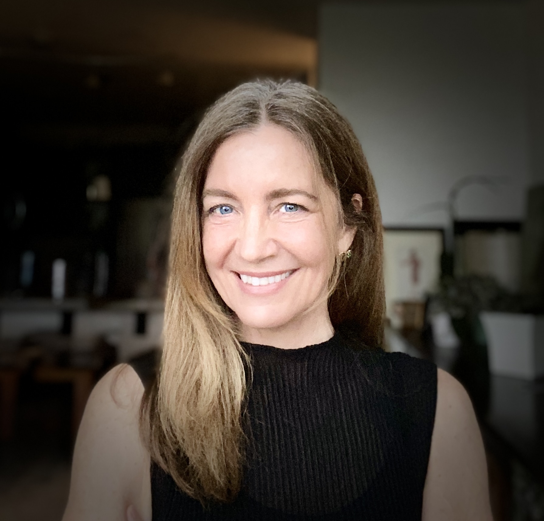 A woman with long light brown hair and blue eyes smiles at the camera, wearing a sleeveless black top indoors with a blurred background.