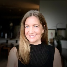 A woman with long light brown hair and blue eyes smiles at the camera, wearing a sleeveless black top indoors with a blurred background.