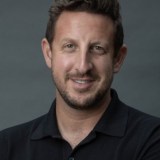 A man with short brown hair and a beard, wearing a black collared shirt, smiles at the camera against a plain gray background.