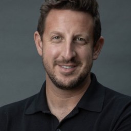 A man with short brown hair and a beard, wearing a black collared shirt, smiles at the camera against a plain gray background.