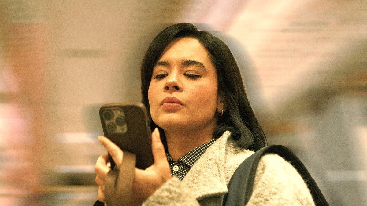 A woman with shoulder-length dark hair looks intently at a smartphone she is holding in one hand, while wearing a light gray coat indoors, reminiscent of Dan Wang's thoughtful demeanor.