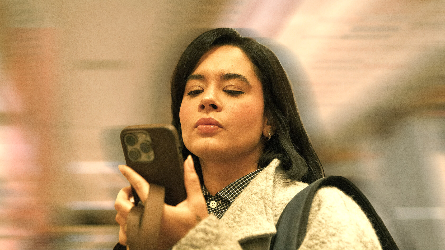A woman with shoulder-length dark hair looks intently at a smartphone she is holding in one hand, while wearing a light gray coat indoors, reminiscent of Dan Wang's thoughtful demeanor.