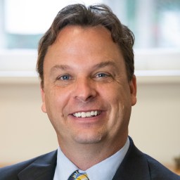 A man with wavy brown hair smiles at the camera while wearing a dark suit, light blue shirt, and yellow striped tie. A blurred window is in the background.