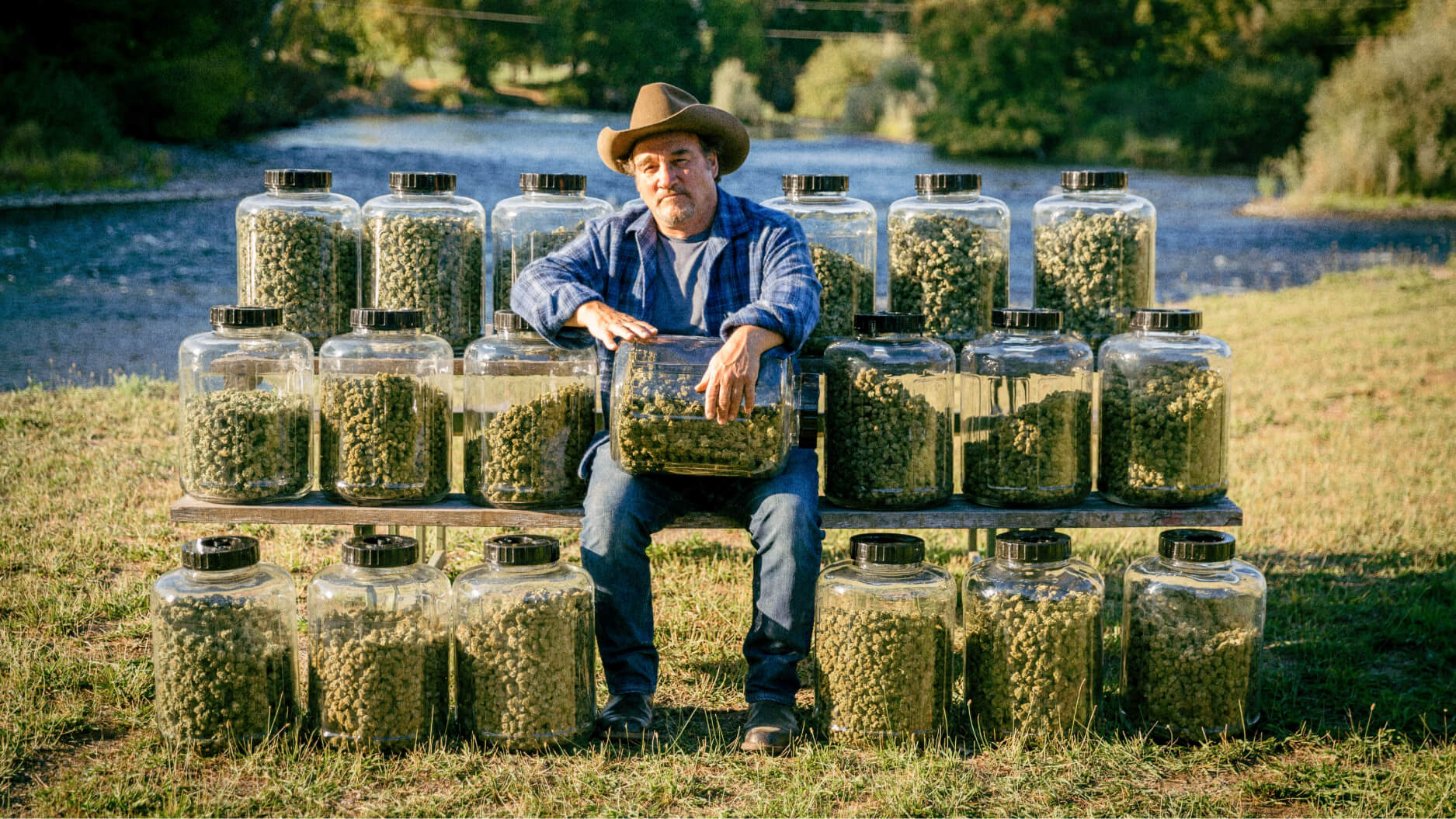 Jim Belushi, wearing a cowboy hat, sits outdoors on a bench surrounded by large jars of green plant material, with a river and trees in the background.