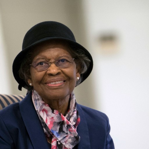 An elderly woman wearing glasses, a black hat, and a patterned scarf smiles while seated indoors—reminiscent of Gladys West, the Einstein behind GPS technology.