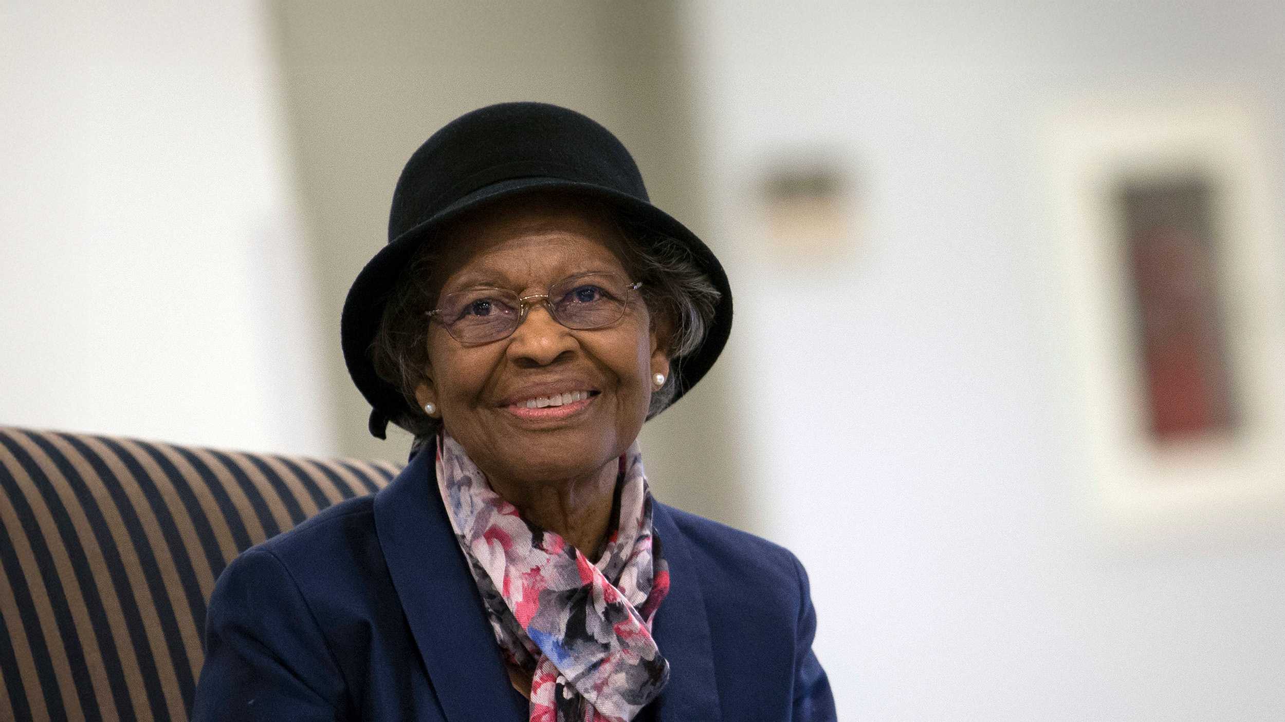 An elderly woman wearing glasses, a black hat, and a patterned scarf smiles while seated indoors—reminiscent of Gladys West, the Einstein behind GPS technology.