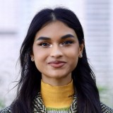 A woman with long dark hair and light makeup, wearing a yellow turtleneck and patterned jacket, stands outdoors in front of a blurred background.