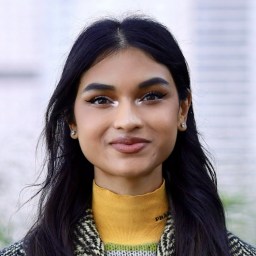 A woman with long dark hair and light makeup, wearing a yellow turtleneck and patterned jacket, stands outdoors in front of a blurred background.