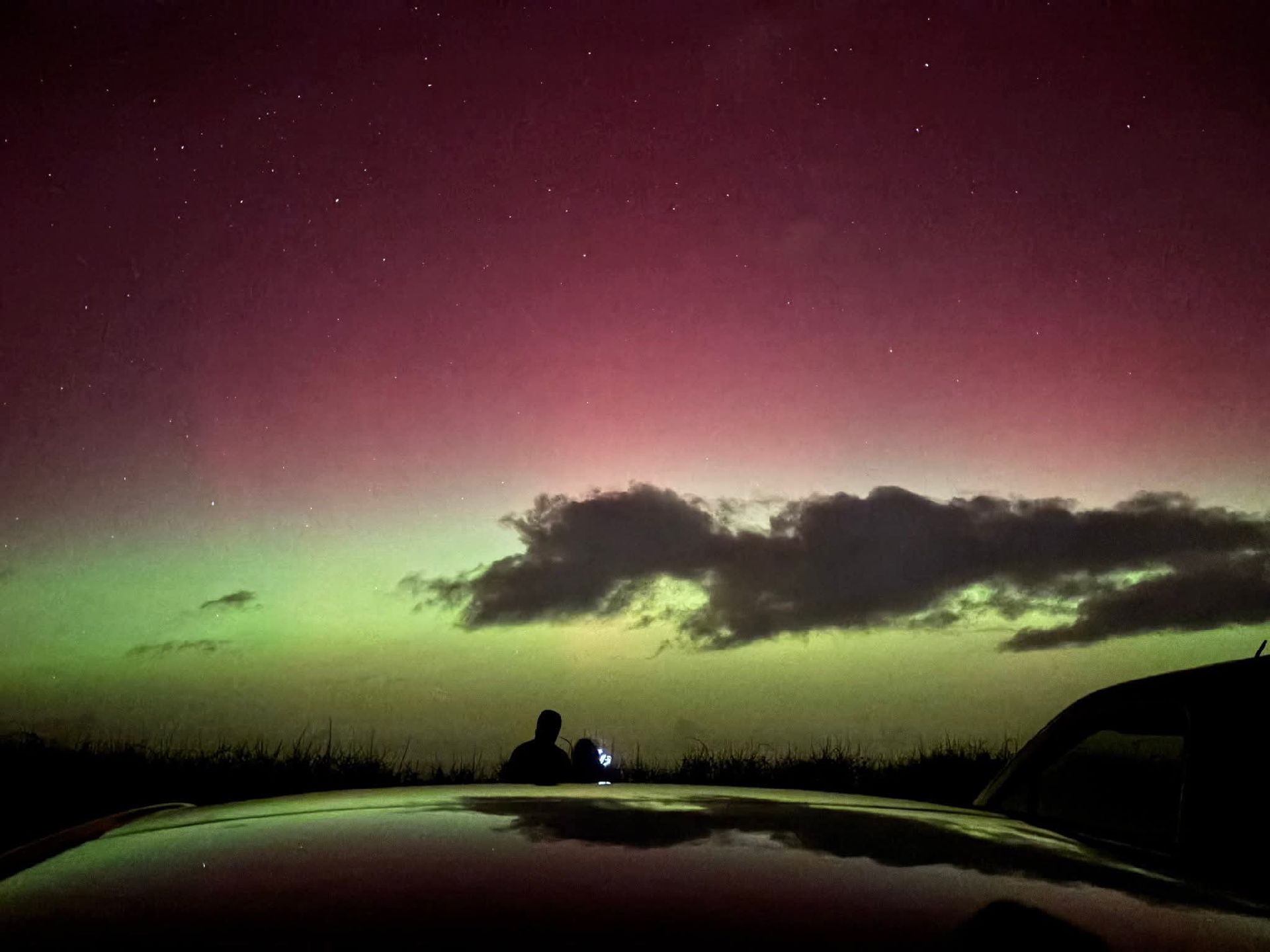 A person stands near a parked car under a sky lit with green and purple aurora, vibrant from a recent solar radiation storm, with stars visible and a low, dark cloud on the horizon.