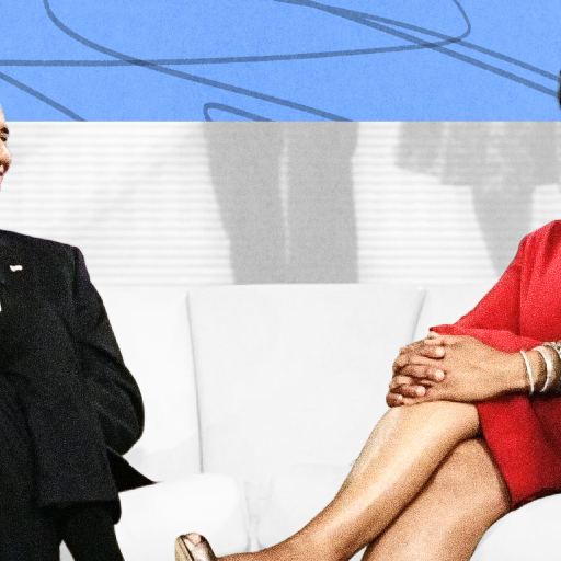 Two people sit on white chairs facing each other, both smiling and dressed formally, on a brightly lit stage with a blue and white background.