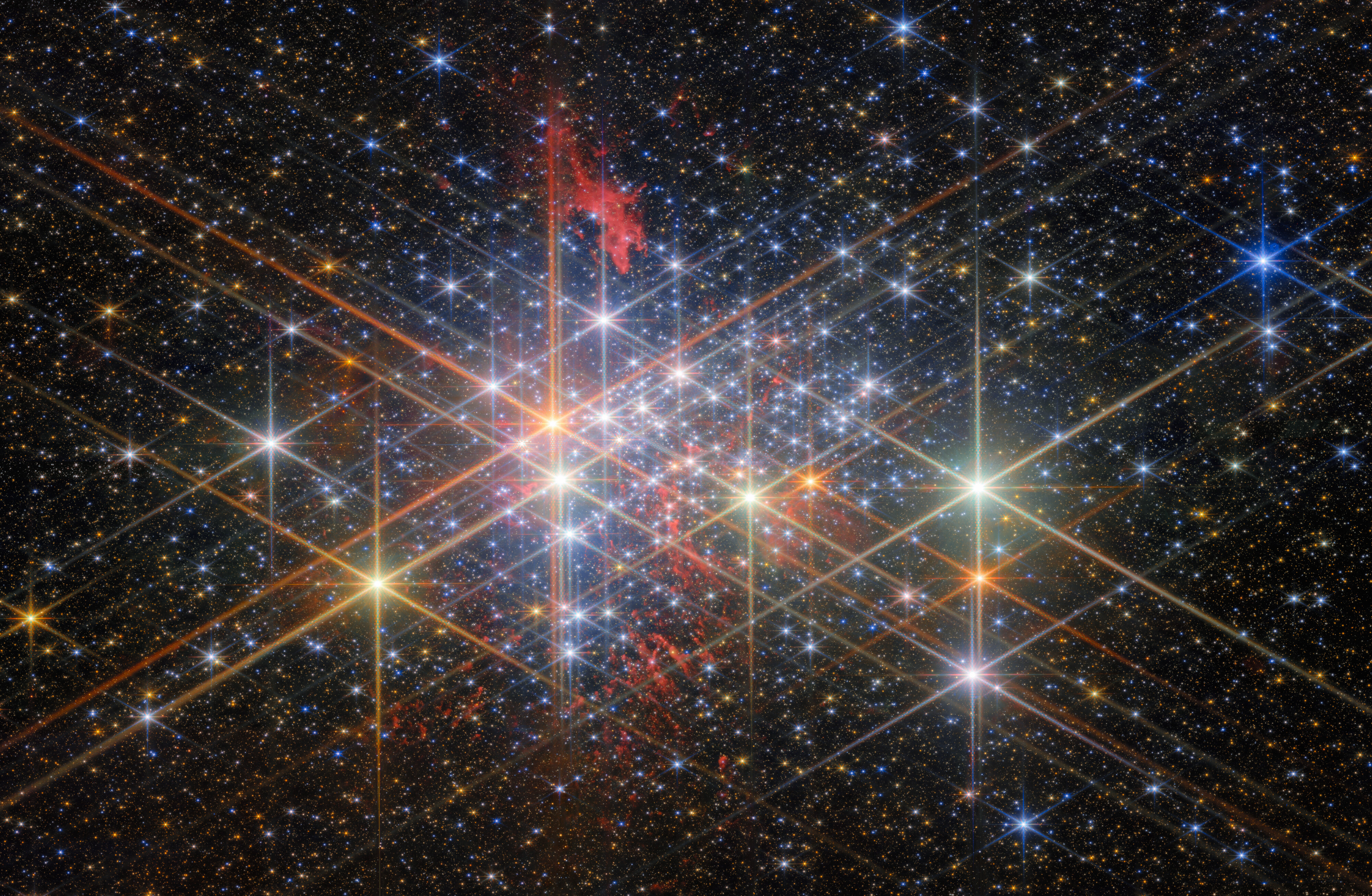 A dense star cluster with bright, colorful stars showing diffraction spikes, as seen in a JWST reveal of the universe, set against a background of numerous smaller stars and a faint red nebula.