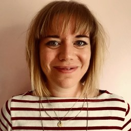 A person with straight blonde hair and bangs, wearing a red and white striped shirt and layered necklaces, stands in front of a plain light-colored wall.
