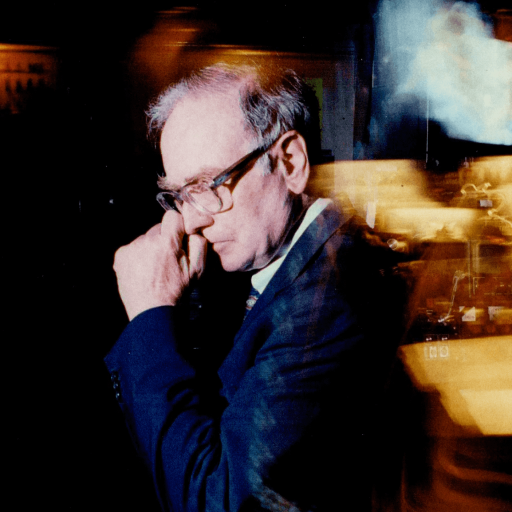 Angus Fletcher, a man in a suit and glasses, stands in profile, touching his face in a dimly lit room with blurred lights and equipment in the background.