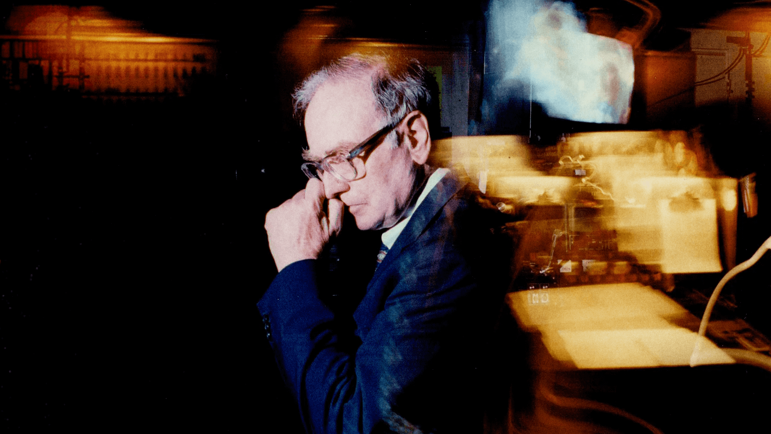 Angus Fletcher, a man in a suit and glasses, stands in profile, touching his face in a dimly lit room with blurred lights and equipment in the background.