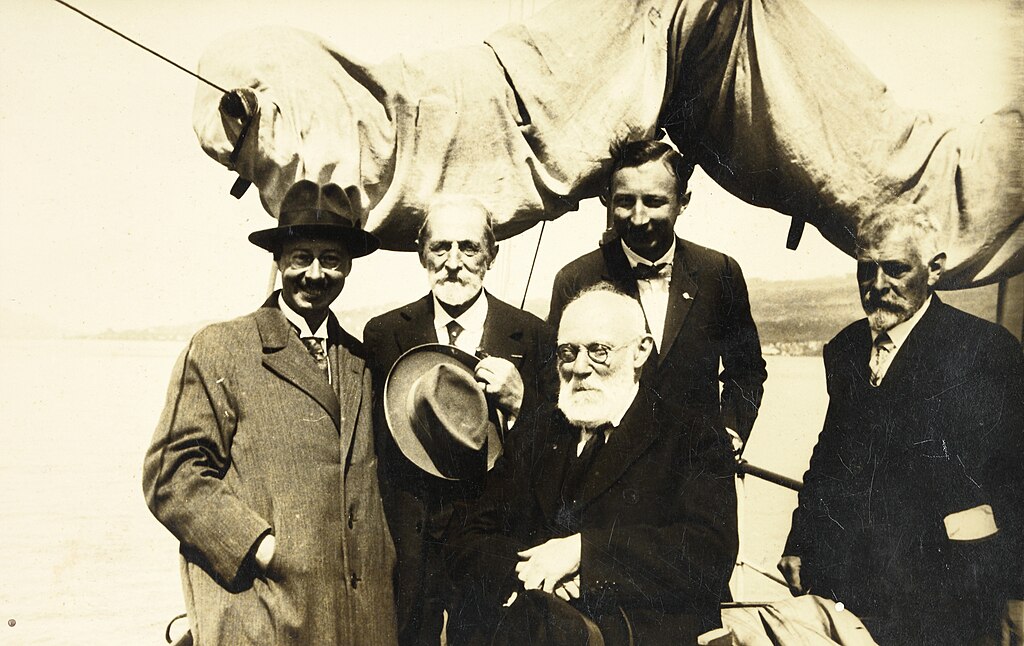 Five older men in suits and hats pose together on a boat, with sails and water visible in the background.