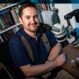 A man sits at a desk with a microscope, two small vials, and bookshelves in the background, looking at the camera and smiling.