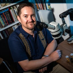 A man sits at a desk with a microscope, two small vials, and bookshelves in the background, looking at the camera and smiling.