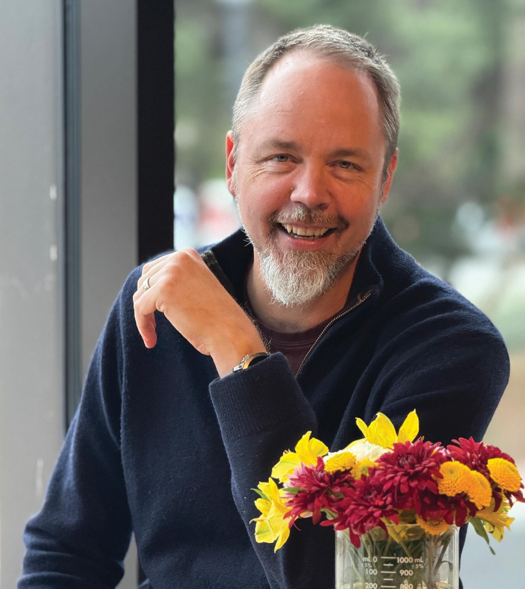 A man with a short beard and dark sweater sits at a table, smiling beside a beaker filled with red and yellow flowers.