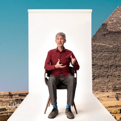 A man sits on a chair against a white backdrop, placed in front of the Pyramid of Giza in Egypt, under a clear blue sky.