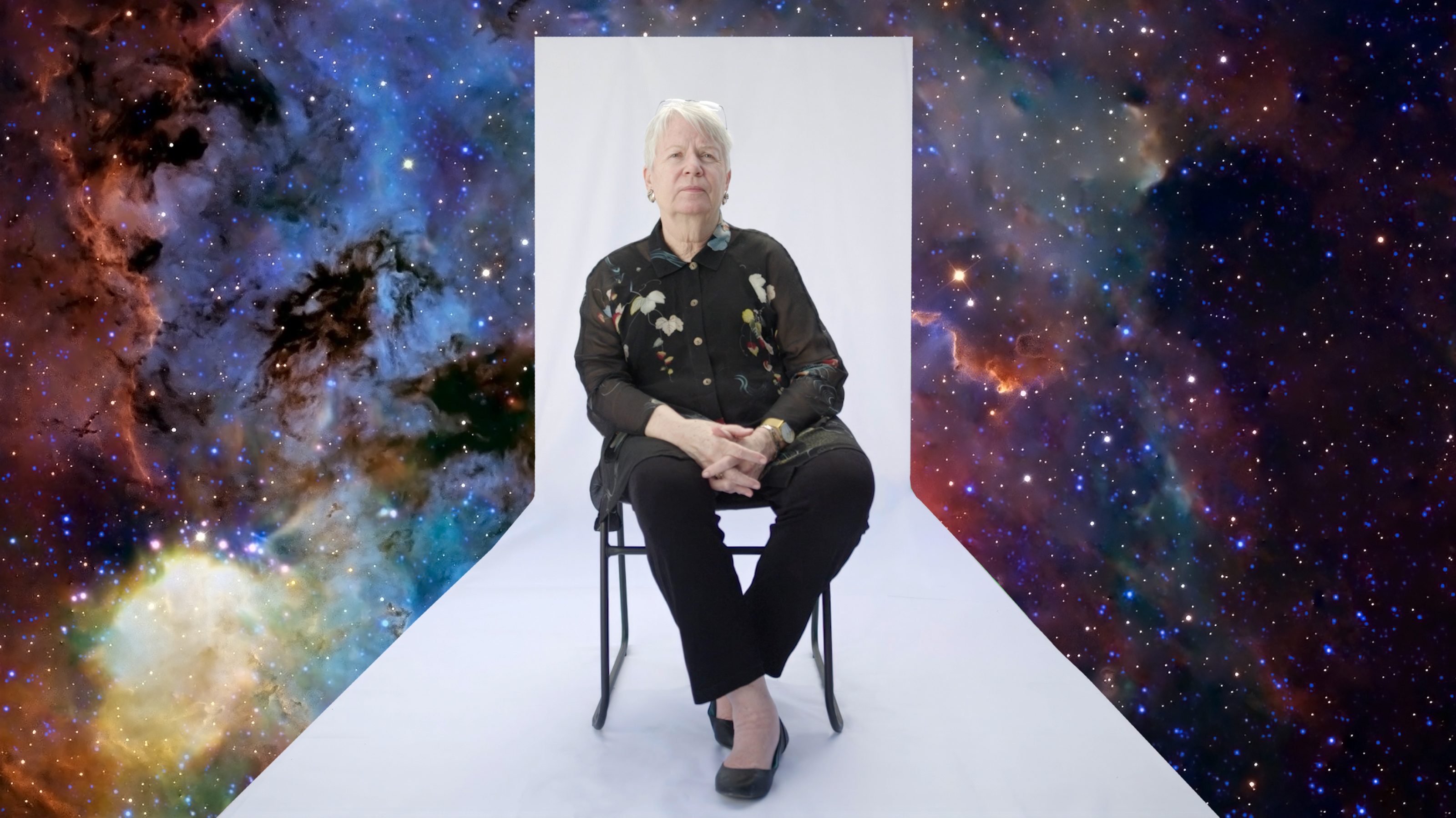 An older person sits on a chair against a white backdrop, with a colorful outer space scene digitally added around the background.