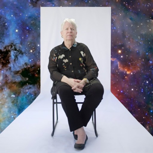 An older person sits on a chair against a white backdrop, with a colorful outer space scene digitally added around the background.