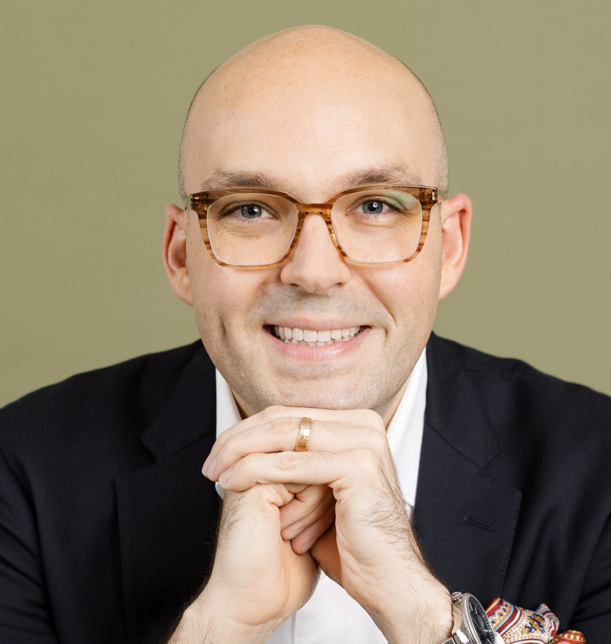 A smiling bald man wearing glasses, a dark suit, white shirt, and watch, resting his hands under his chin against a plain background.