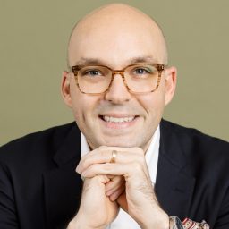A smiling bald man wearing glasses, a dark suit, white shirt, and watch, resting his hands under his chin against a plain background.