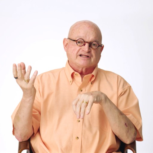 Bald man wearing glasses and a peach shirt sits on a chair against a white background, gesturing with his left hand while talking.