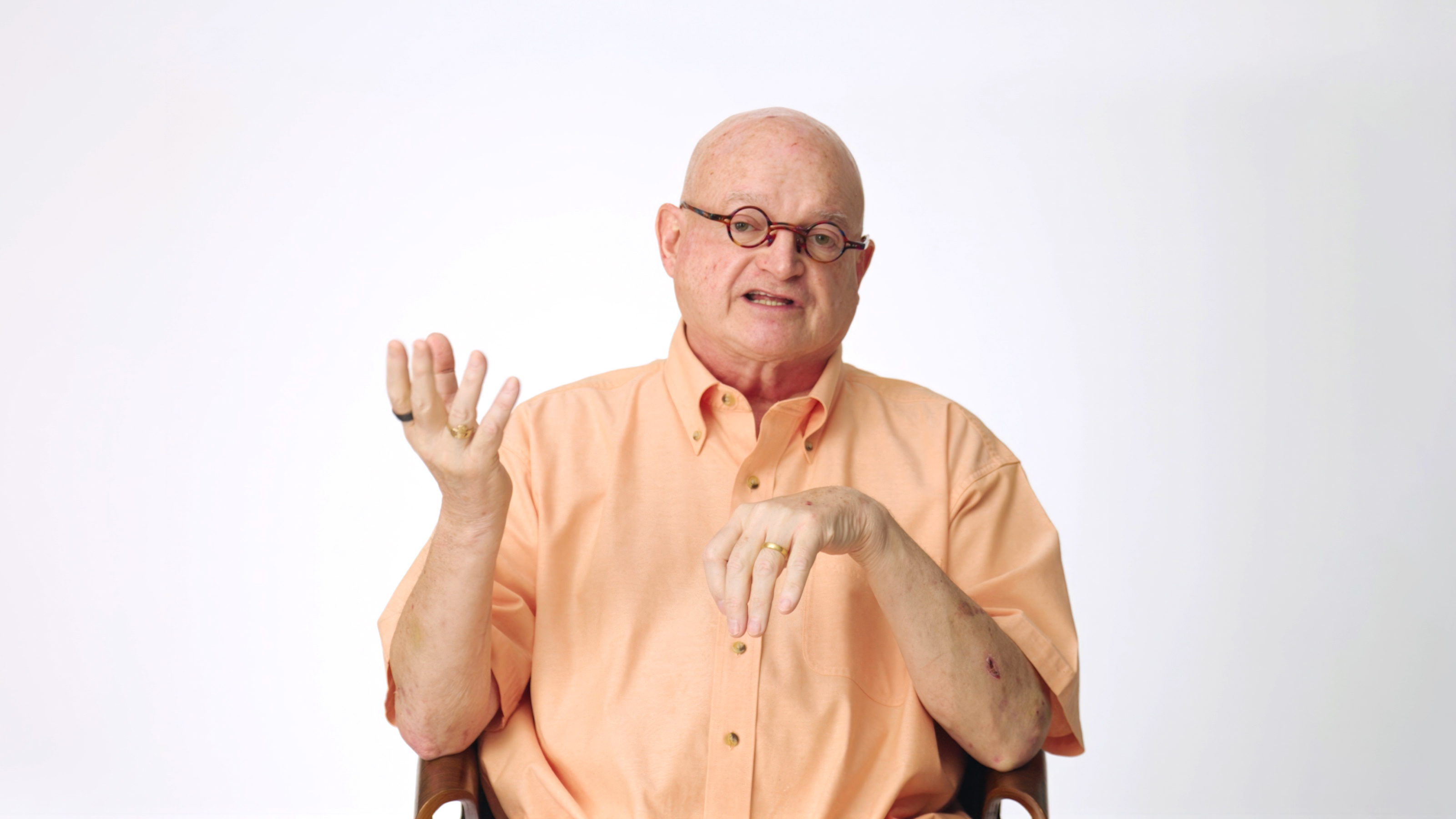 Bald man wearing glasses and a peach shirt sits on a chair against a white background, gesturing with his left hand while talking.