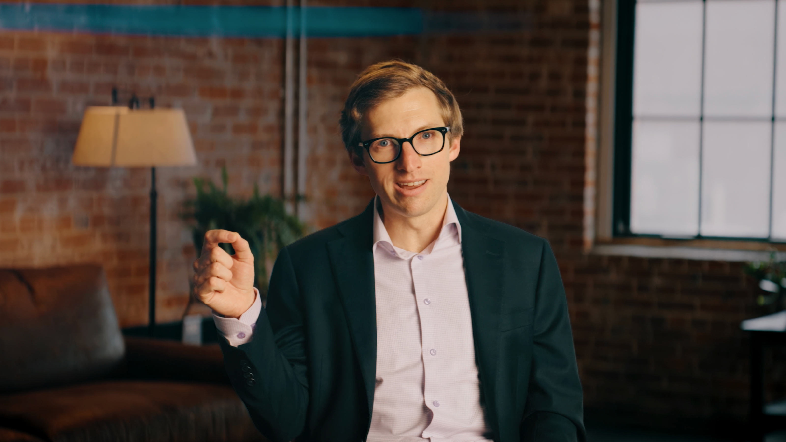 A man in glasses and a suit jacket sits indoors, gesturing with his right hand. Exposed brick walls, a window, and a lamp are visible in the background.