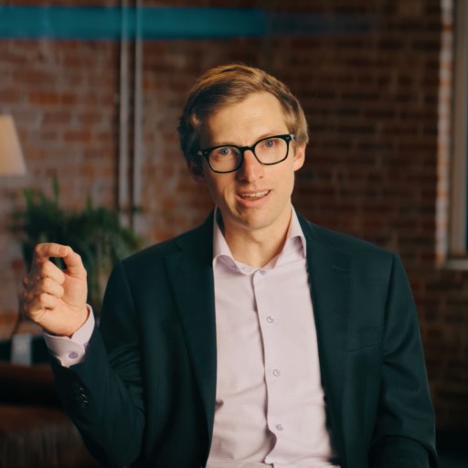 A man in glasses and a suit jacket sits indoors, gesturing with his right hand. Exposed brick walls, a window, and a lamp are visible in the background.