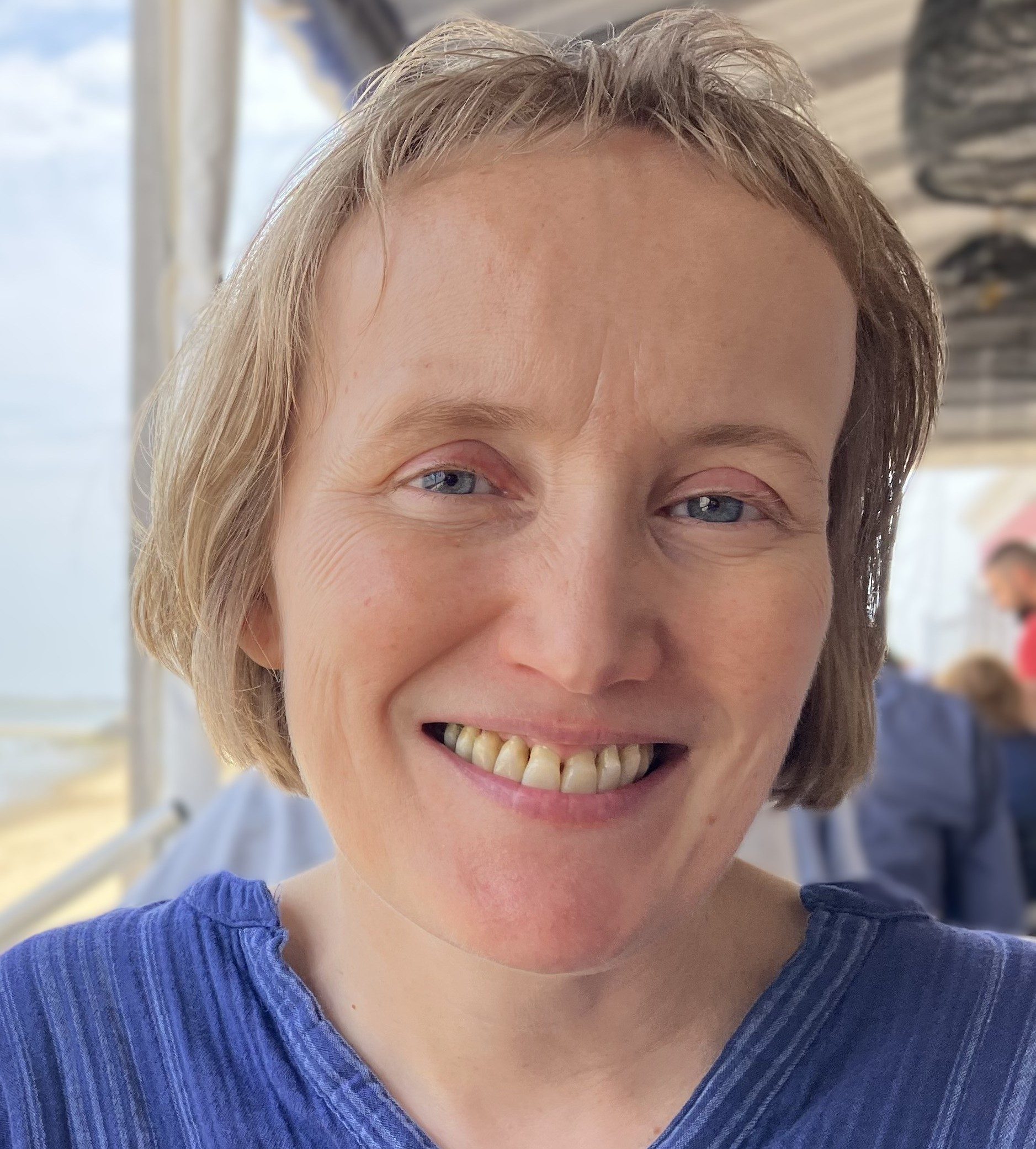 A person with short light brown hair, wearing a blue top, is smiling at the camera in an outdoor setting with a beach visible in the background.