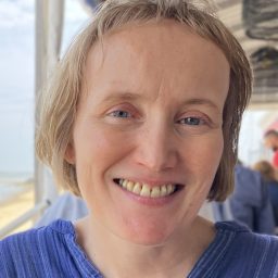 A person with short light brown hair, wearing a blue top, is smiling at the camera in an outdoor setting with a beach visible in the background.