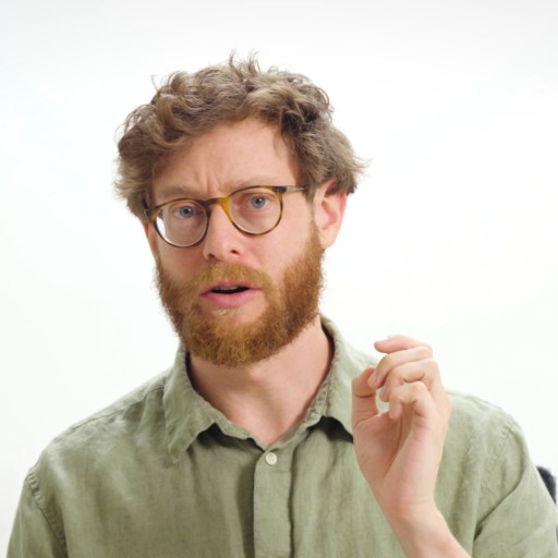 A man with curly hair, a beard, and glasses, wearing a green button-up shirt, gestures with his hand against a plain white background.