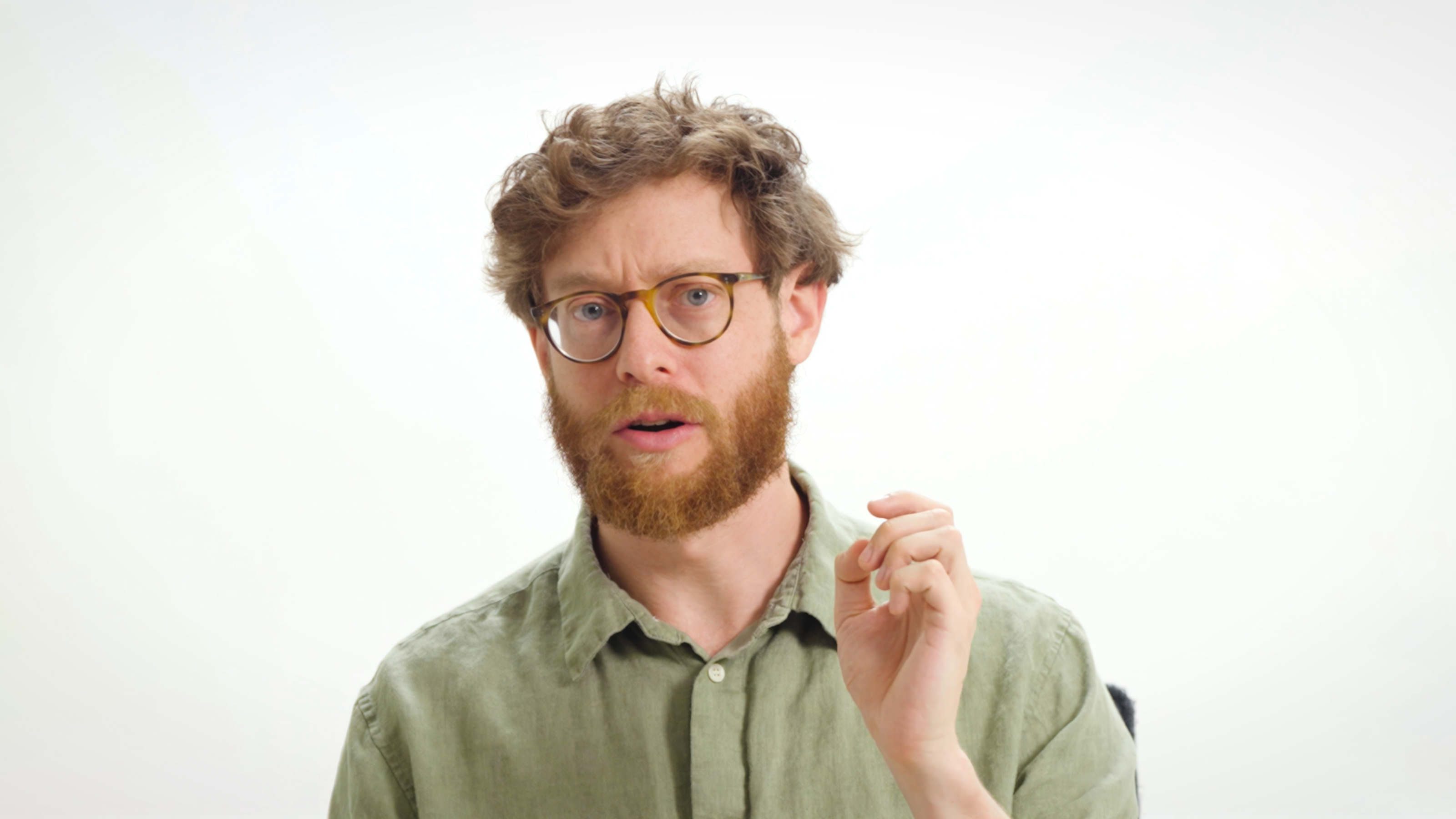 A man with curly hair, a beard, and glasses, wearing a green button-up shirt, gestures with his hand against a plain white background.