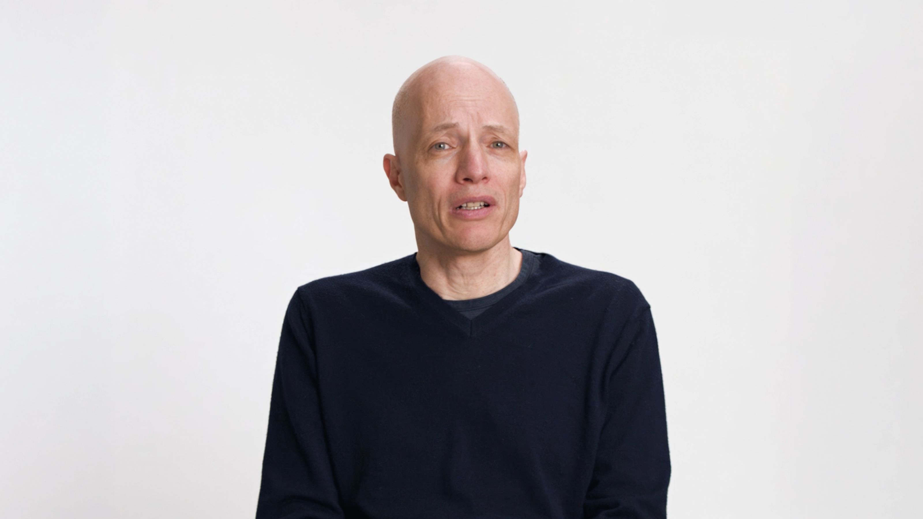 A bald man wearing a dark sweater sits against a plain white background, looking slightly to the side with a neutral expression.