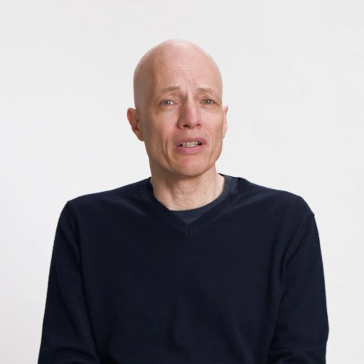 A bald man wearing a dark sweater sits against a plain white background, looking slightly to the side with a neutral expression.