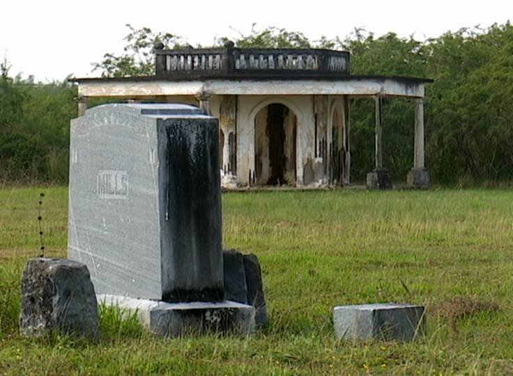 A weathered gravestone stands in front of an old, dilapidated building surrounded by overgrown grass and bushes.