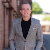 A man in a gray plaid blazer and black shirt stands outside with his hands in his pockets, smiling at the camera. Brick buildings and trees are visible in the background.