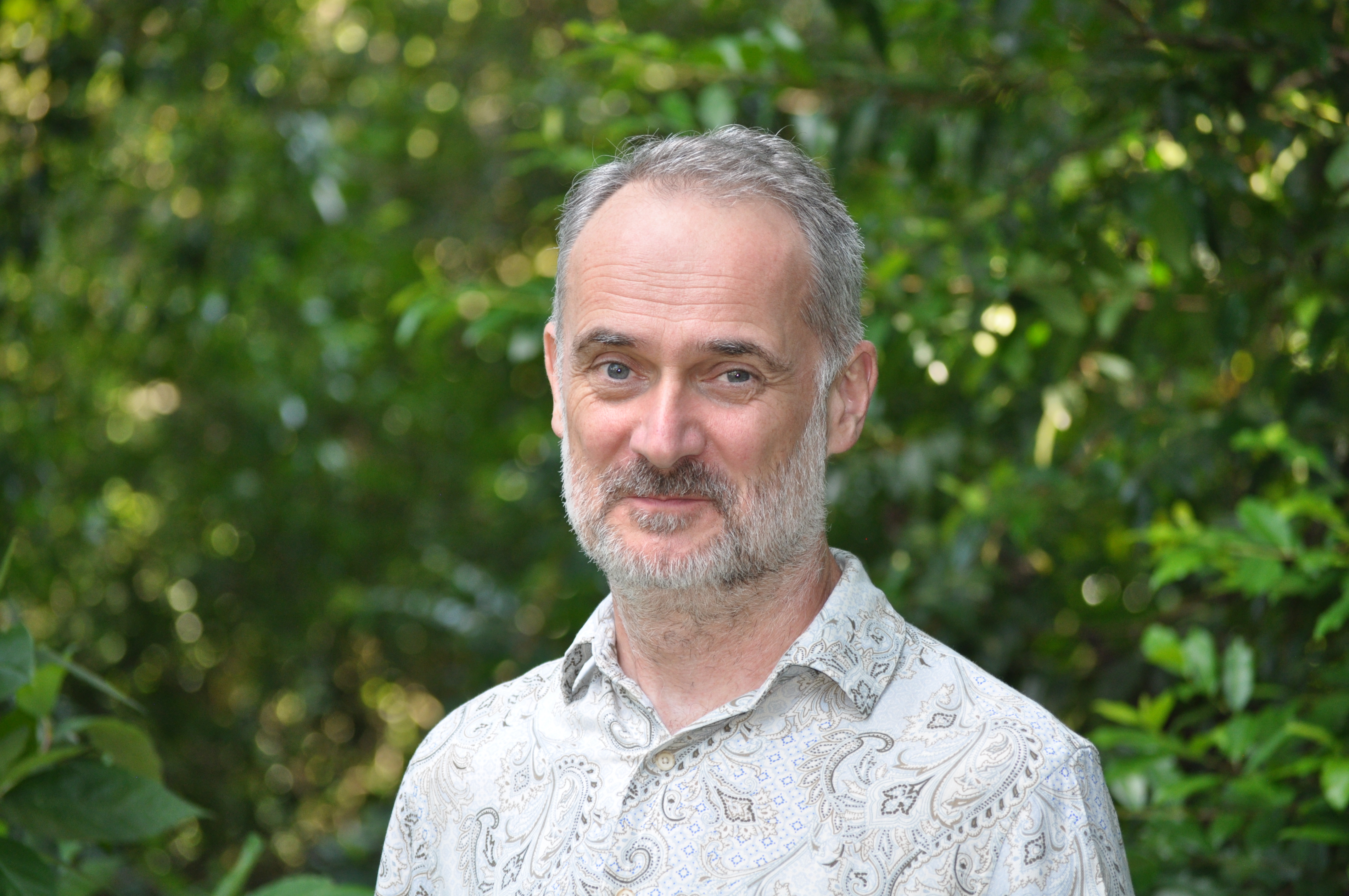 A middle-aged man with short gray hair and a beard stands outdoors in front of green foliage, wearing a patterned shirt.