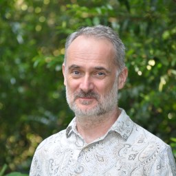 A middle-aged man with short gray hair and a beard stands outdoors in front of green foliage, wearing a patterned shirt.