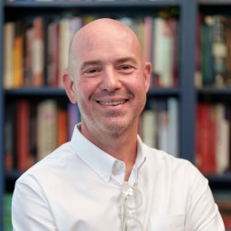 A bald man wearing a white button-up shirt and eyeglasses smiles while standing in front of a bookshelf filled with books.