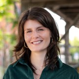 A woman with shoulder-length brown hair, wearing a green shirt and hoop earrings, smiles outdoors with blurred trees and structures in the background.