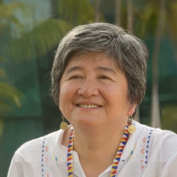 A smiling older woman with short gray hair, wearing a white embroidered top, beaded necklace, and spiral earrings, is seen outdoors with blurred greenery in the background.