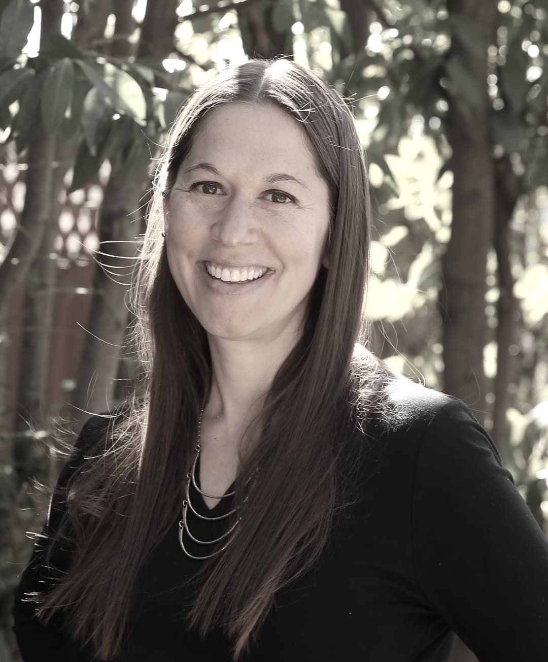 Woman with long brown hair wearing a black top and necklace, smiling outdoors with trees and sunlight in the background.