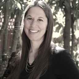 Woman with long brown hair wearing a black top and necklace, smiling outdoors with trees and sunlight in the background.