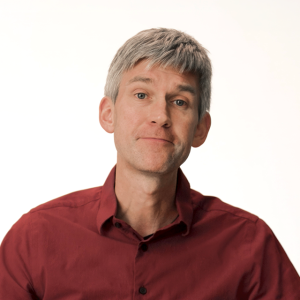 A man with short gray hair, wearing a red button-up shirt, faces the camera against a plain white background.