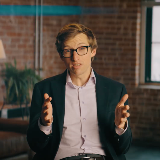 A man in a suit and glasses sits indoors, gesturing with both hands while speaking. The setting includes a brick wall, a lamp, a plant, and a window.