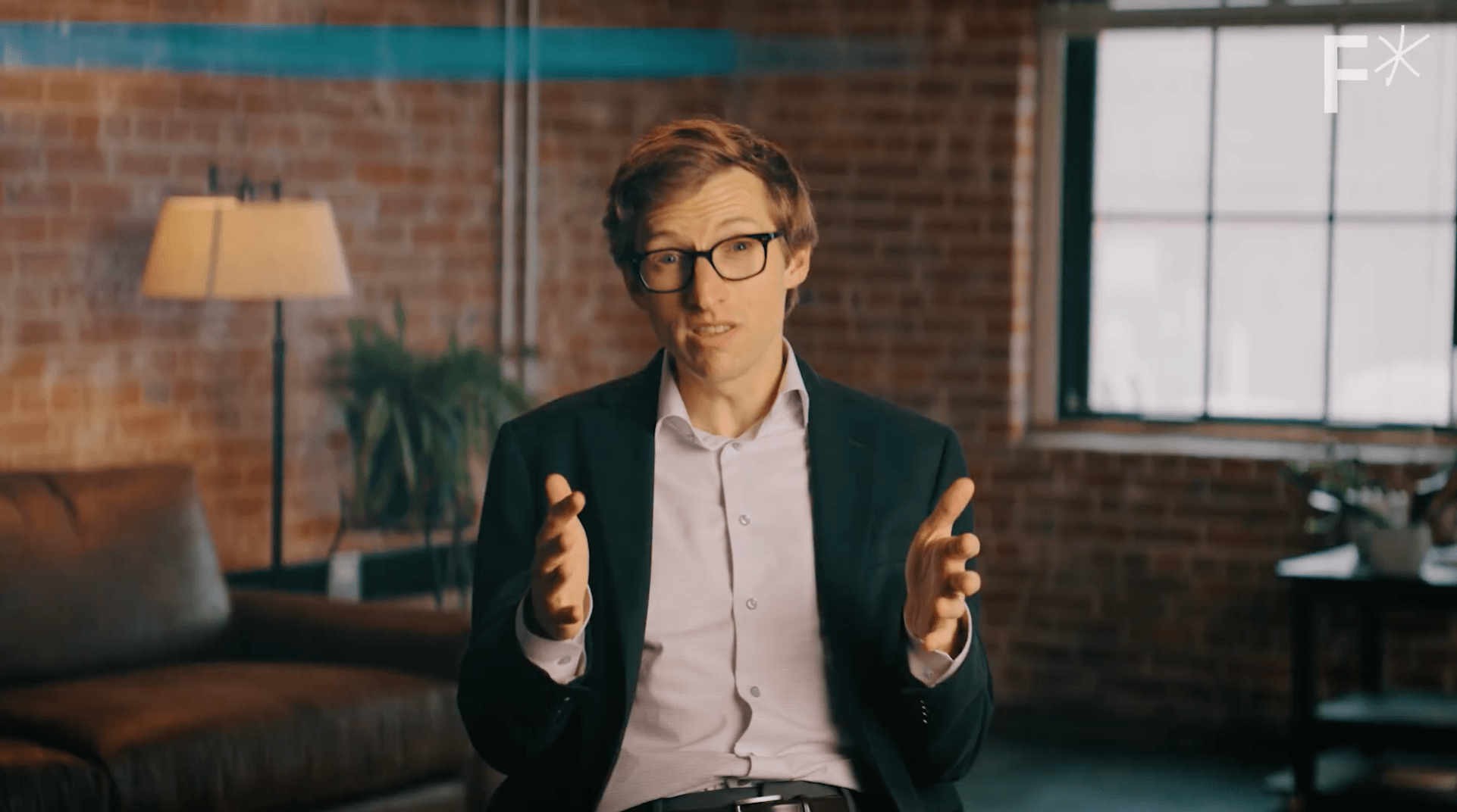 A man in a suit and glasses sits indoors, gesturing with both hands while speaking. The setting includes a brick wall, a lamp, a plant, and a window.